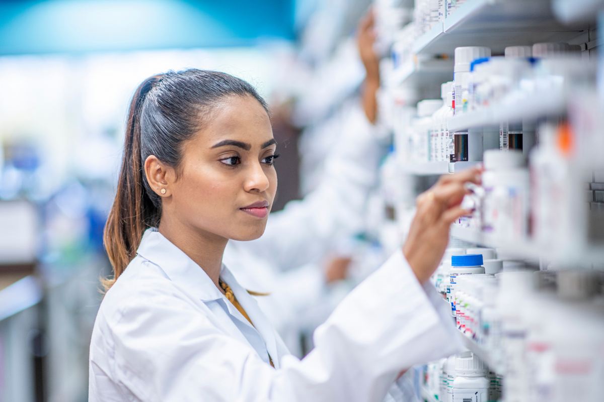 Pharmacist in white coat selects a bottle from pharmacy shelf filled with various medicines. Background is brightly lit. Focused expression.