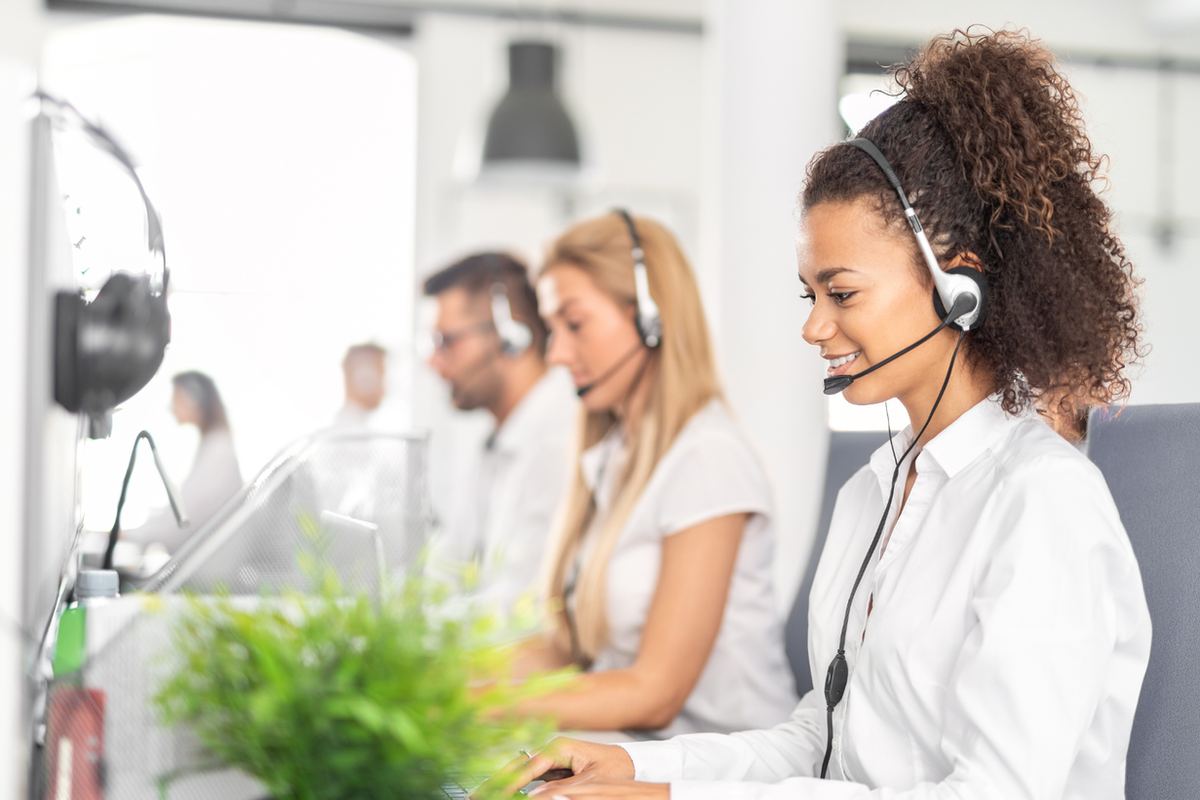 Smiling headset-wearing woman works at a computer in a bright call center, with coworkers blurred in the background.