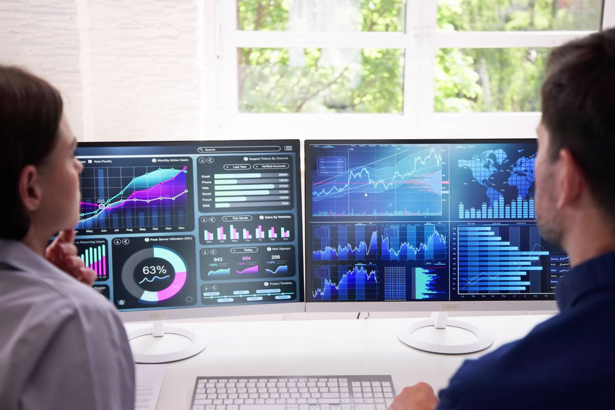Two people analyzing data on dual monitors displaying charts and graphs. Bright office with greenery outside the window creates a focused mood.