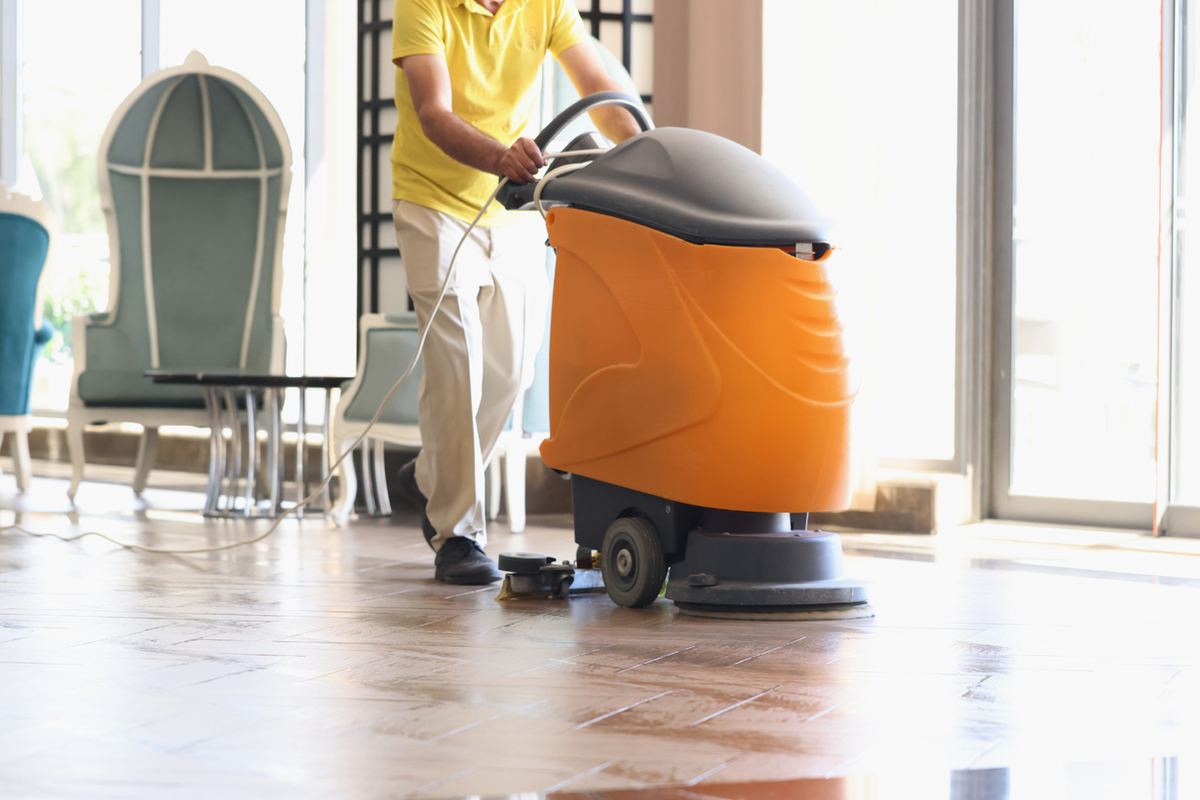 Person in yellow shirt operating an orange floor cleaner in a brightly lit room with blue chairs and large windows.
