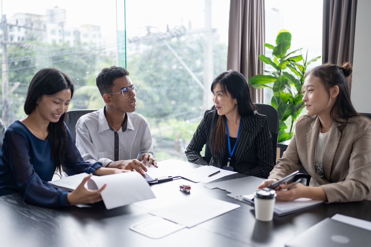 Four coworkers discuss papers around a table in a bright office with large windows, a potted plant, and relaxed smiles.